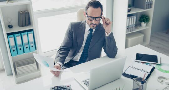 ERP Budget uomo con occhiali in giacca e cravatta seduto alla scrivania che guarda il portatile mentre ha un foglio in mano