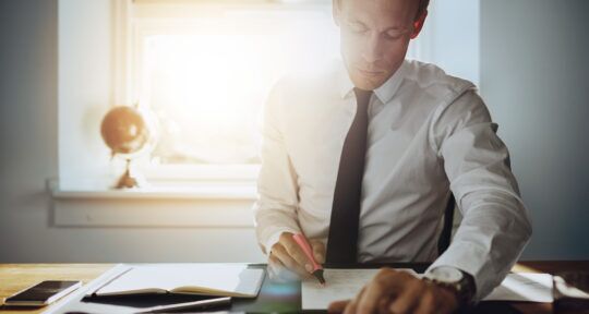 blog ERP Financial Management uomo in giacca e cravatta seduto alla scrivania e che scrive su un foglio